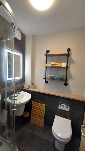 Un baño con cabina de ducha de vidrio, un lavabo blanco, un inodoro y una repisa con toallas y artículos de aseo.