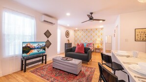 Una sala de estar con televisor, sofá, reposapiés y un comedor con mesa y sillas.