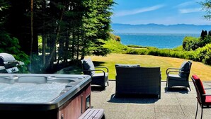Une terrasse dotée d’un jacuzzi, de fauteuils en osier et d’une table avec des chaises rouges, offrant une vue sur une pelouse et la mer.