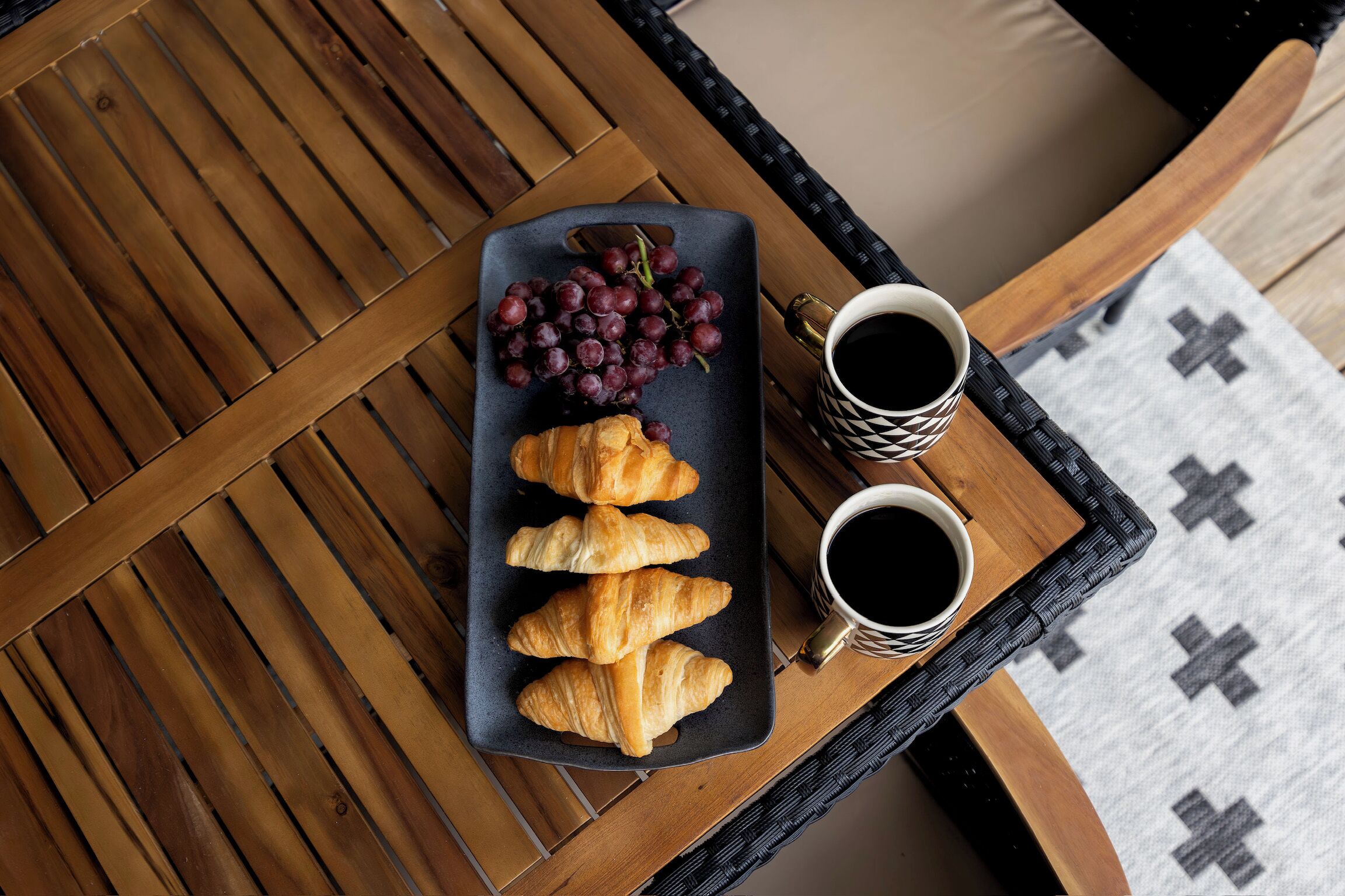 Une table en bois sur laquelle se trouve une assiette noire contenant des croissants et des raisins, ainsi que deux tasses de café.