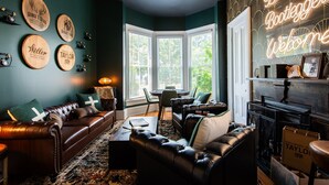 Ein gemütliches Wohnzimmer mit einer dunkelgrünen Wand, Ledersofas, einem Couchtisch und einem Kamin. An der Wand befinden sich gerahmte Gegenstände und ein Neonzeichen.