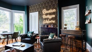 Ein gemütliches Wohnzimmer mit einem Klavier, Ledersofas, einem Couchtisch und einem Neonlichtschild mit der Aufschrift "Bourbon Codeggers Welcome".
