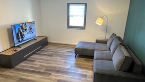 A modern living room with a flat-screen TV, a grey sectional sofa, a wooden floor, and a window with blinds.
