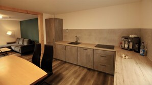 A modern kitchen with a wooden countertop, a sink, and a coffee machine. There is a dining table with chairs and a sofa in the adjacent living area.