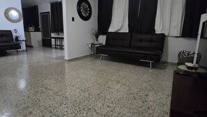 A spacious living area with a ceiling fan, a dark-colored sofa, a small table with a plant, and a kitchen area in the background.