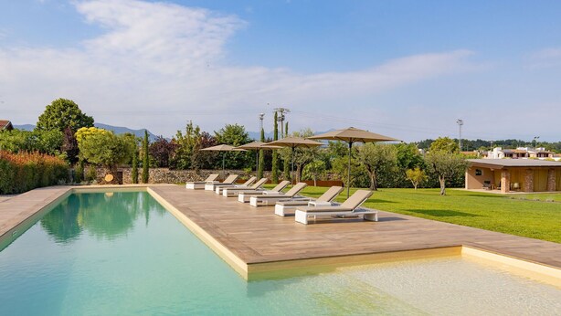 A pool area with lounge chairs and umbrellas, surrounded by greenery and a clear sky.