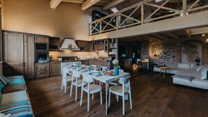A spacious loft-style living area with a dining table, white chairs, and a high ceiling with exposed wooden beams.