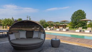 A wicker lounge chair with cushions by a poolside, a round table with a glass, and lounge chairs with umbrellas in the background.