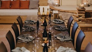 A dining table set for six with wine bottles, glasses, and folded napkins. The room features a stone wall and a wooden door.