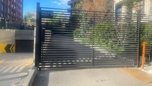 Une entrée fermée par une barrière métallique, une route en béton avec des marquages jaunes et, au loin, des immeubles à plusieurs étages.
