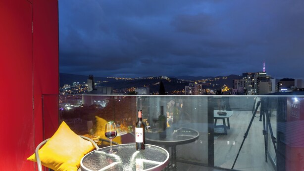 Un balcon avec une table en verre, une bouteille de vin et un coussin jaune.