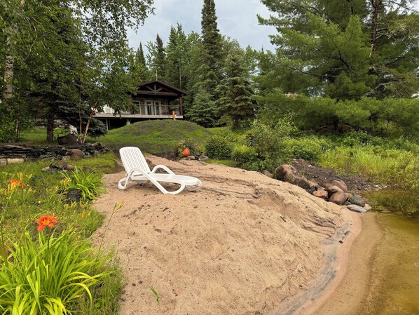 A sandy area with a white lounge chair, surrounded by greenery and a house in the background. - Amazing sunsets and fishing 🎣 on this lakefront cabin just minutes from Kenora! (Kenora)