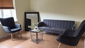 En stue med en grå sofa, to sorte lænestole, et rundt bord med en vase og et stort spejl på væggen.