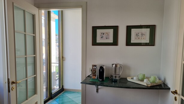 A hallway with a glass door leading to a balcony, a small shelf with a kettle and cups, and two framed pictures on the wall.