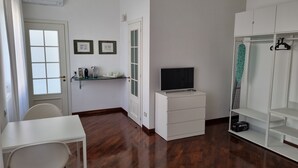 A room with a wooden floor, a white desk with a chair, a TV on a white cabinet, a wardrobe, and a door with a window.