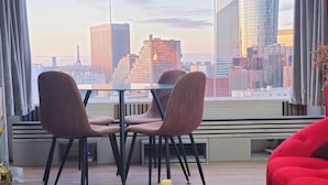 A modern room with a large window offering a city view, including a tall building and the Eiffel Tower. There is a dining table with chairs, a red sofa, and a small plant on the windowsill. - Charming 2-bedroom apartment with big Terrace , view of Eiffel Tower and Paris (Courbevoie)