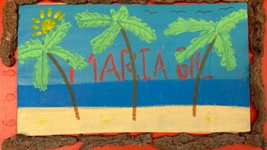 Une scène de plage avec des palmiers et le nom « Maria G. » peint sur une planche de bois.