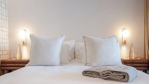 A neatly made bed with white linens, two bedside tables with bottles and glasses, and wall-mounted light fixtures. - Seaside 8: Stylish Seaside Studio with Balcony (Cape Town)