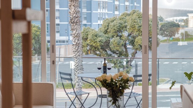 A modern living room with a glass door leading to a balcony, a round wooden table with a vase of flowers, and a view of a cityscape and water. - Seaside 8: Stylish Seaside Studio with Balcony (Cape Town)