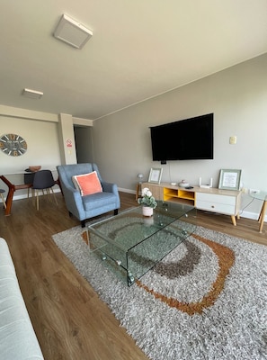 Una sala de estar moderna con una mesa de centro de vidrio, un sillón azul, un televisor de pantalla plana montado en la pared y piso de madera.