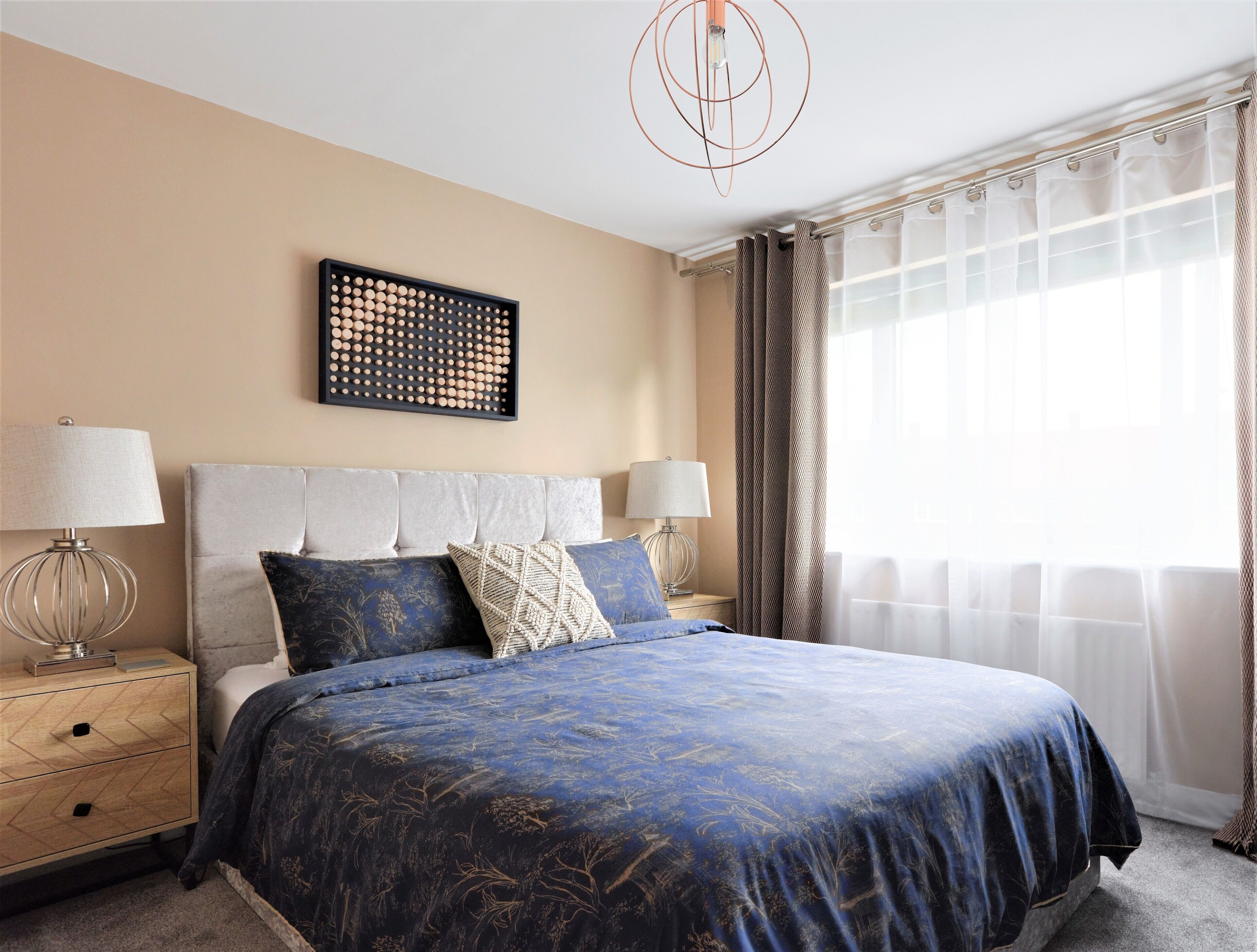 A bedroom with a large bed, two bedside tables with lamps, a wall-mounted artwork, and a window with curtains.