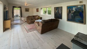 Un ampio soggiorno con un divano marrone, un tavolino da caffè e una televisione. Sono presenti due finestre con tende, un quadro appeso al muro e un mobiletto in legno.
