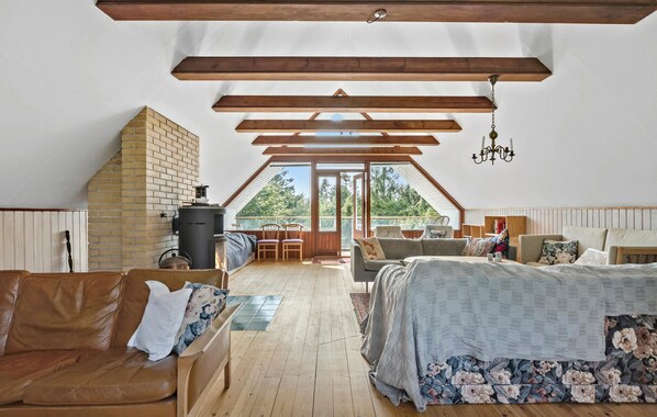 An attic bedroom with a sloped ceiling, wooden beams, a brick fireplace, a bed with floral bedding, a sofa, and a view of greenery outside. - Stunning home in Hundested with WiFi (Hundested)