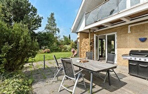 An outdoor patio with a wooden table and chairs, a grill, and a garden with trees and flowers. - Stunning home in Hundested with WiFi (Hundested)