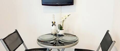 A room with a round glass table, two foldable chairs, a flat-screen TV mounted on the wall, and a small vase with flowers.