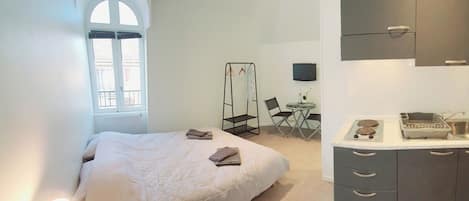 A bedroom with a bed, a bedside table with a lamp, a window with a view, a small dining area with a table and chairs, and a kitchenette with cabinets and a sink.