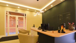 Una recepción con un ordenador, una máquina de café y objetos decorativos. Hay una silla frente al mostrador y una puerta con un cartel encima.