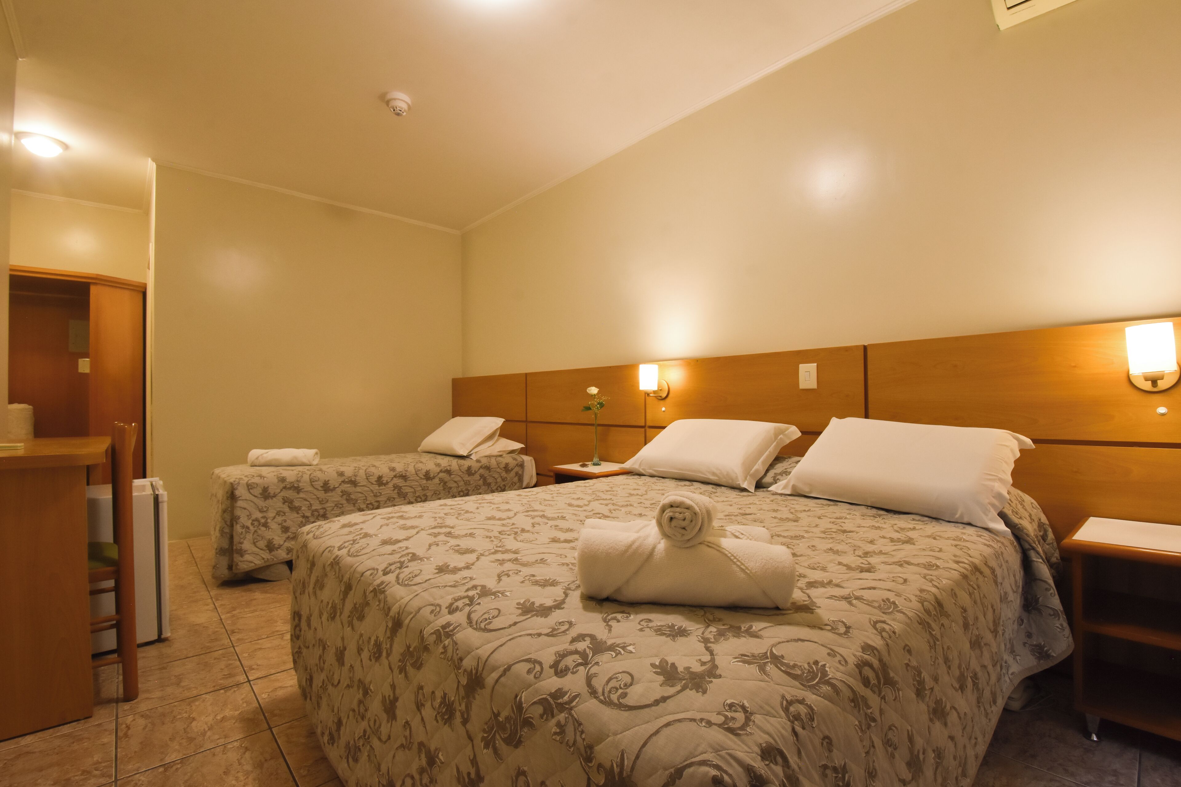 A hotel room with two beds, a wooden headboard, bedside lamps, a small table, and a chair.