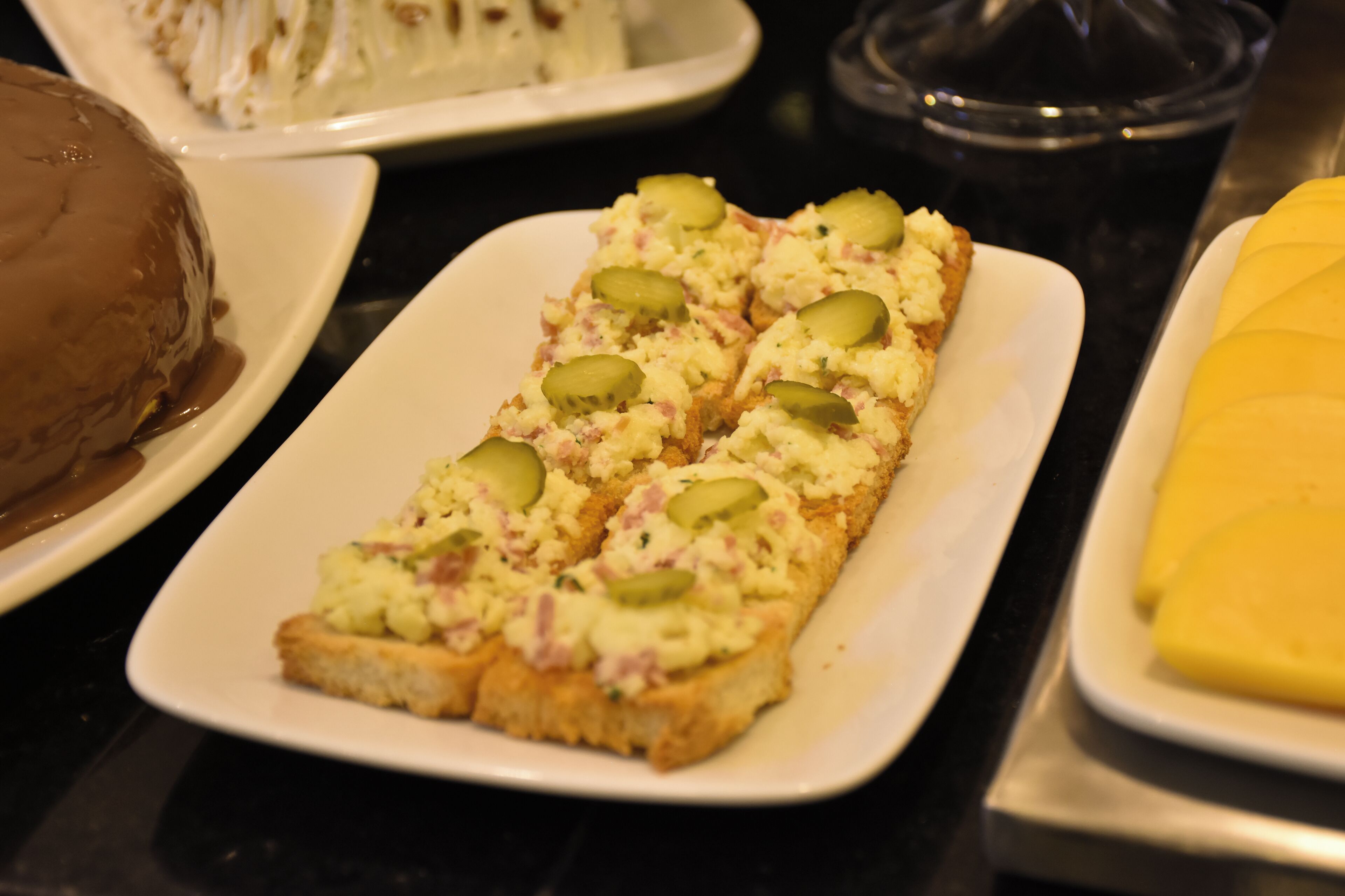 Un plato rectangular blanco con un guiso en capas, posiblemente una entrada o aperitivo, cubierto con pepinillos y una crema.