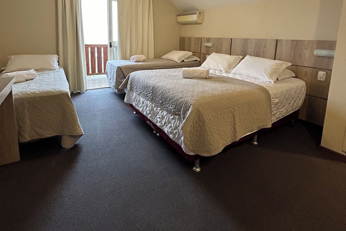 A hotel room with two single beds, a large bed, a ceiling fan, an air conditioning unit, and a balcony with curtains.