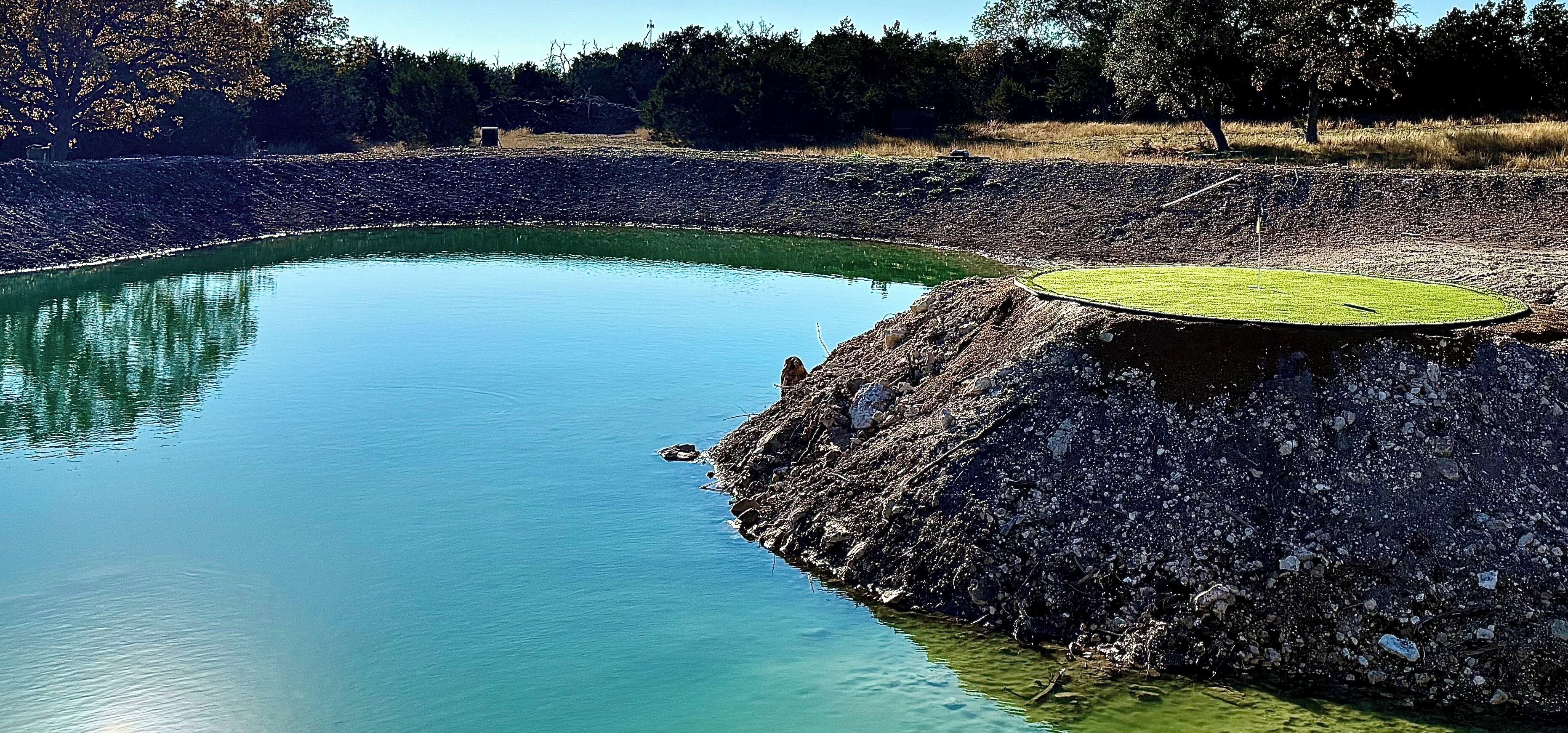 A large, circular pond with a grassy area in the center, surrounded by a rocky embankment.