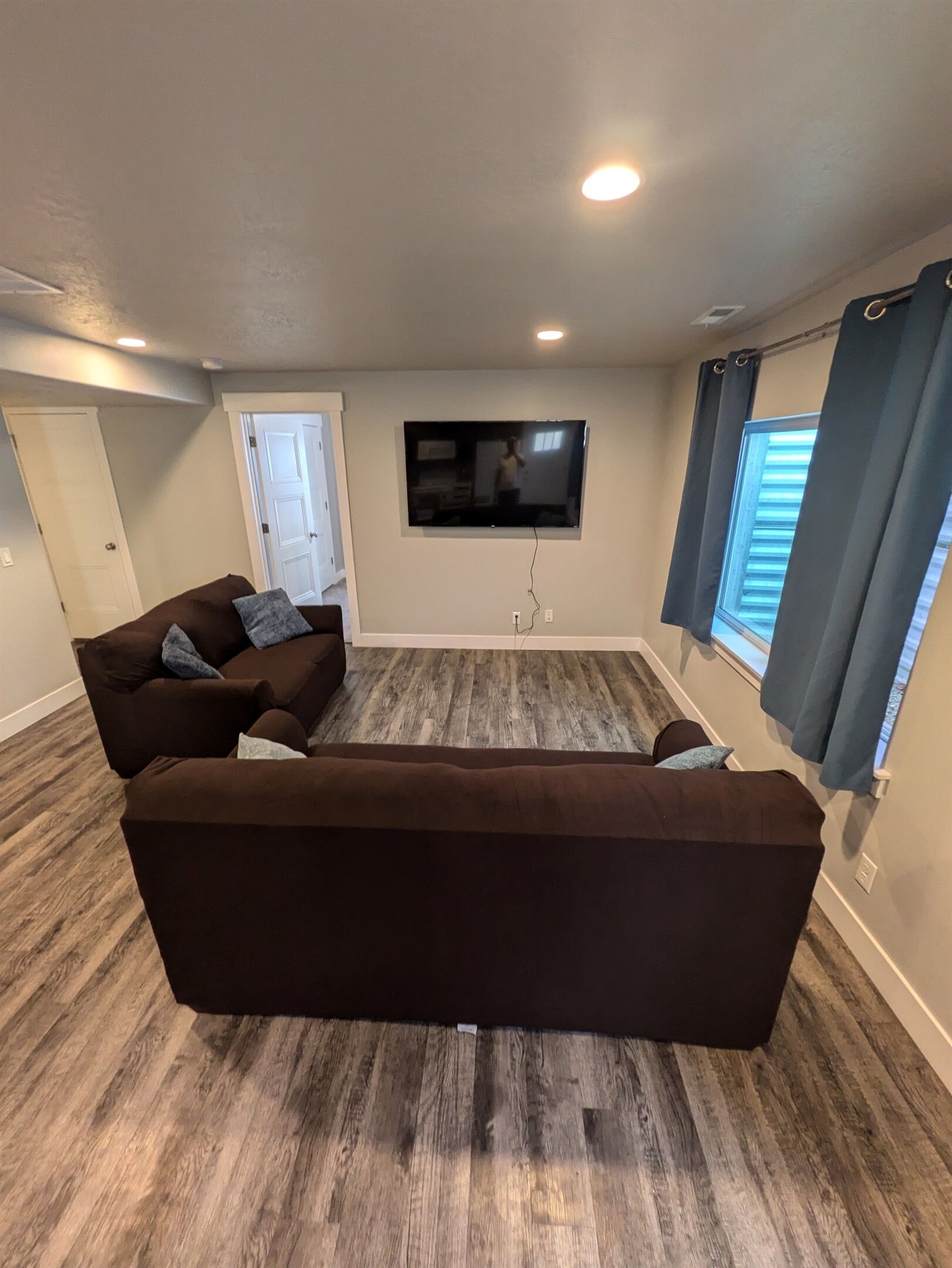 A living room with a large brown sofa, a flat-screen TV mounted on the wall, and a wooden floor.