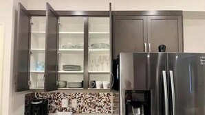 Una cocina con refrigerador de acero inoxidable, armarios abiertos con vajilla, cafetera y salpicadero de azulejos.