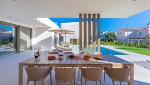 A modern outdoor dining area with a pool, featuring a wooden table, wicker chairs, and a clear view of the surrounding landscape.