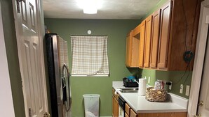 A narrow kitchen with wooden cabinets, a refrigerator, and a trash bin. - Genroc-"It's your home too" (Tallahassee)