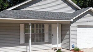 A single-story house with a grey siding exterior, a front porch with white trim, a garage door, and a small landscaped area with mulch and shrubs. - Genroc-"It's your home too" (Tallahassee)