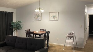 A living room with a dark-colored sofa, a dining table with chairs, a baby high chair, and a ceiling fan. - Genroc-"It's your home too" (Tallahassee)