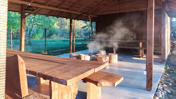 Un espace extérieur abrité, doté d’une table et de bancs en bois, et entouré d’un cadre naturel avec des arbres et une clôture.