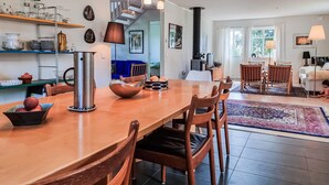 Una cocina moderna con una mesa de madera, sillas y una cafetera. Hay un rincón comedor con alfombra y un televisor al fondo.