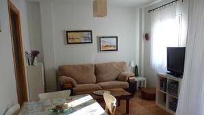Una sala de estar con un sofá, un televisor sobre un mueble bajo, una mesa de comedor con sillas y dos cuadros en la pared.