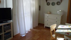 Una habitación con una televisión sobre un mueble bajo, una mesa de comedor con sillas, un armario blanco y un conjunto de platos decorativos en la pared.