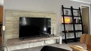 Uma sala de estar com lareira de pedra, uma televisão de tela plana fixada nela, uma estante preta e uma cadeira de madeira.