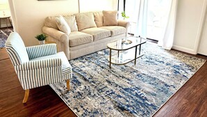 Uma sala de estar com uma poltrona listrada, um sofá bege, uma mesa de centro de vidro e um tapete estampado.