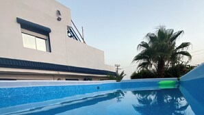 A swimming pool with clear blue water, a white building with a window, and a palm tree in the background.