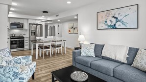 Un salon moderne avec un canapé gris, un fauteuil bleu à motifs, une table basse noire et un coin repas comprenant une table et des chaises blanches.