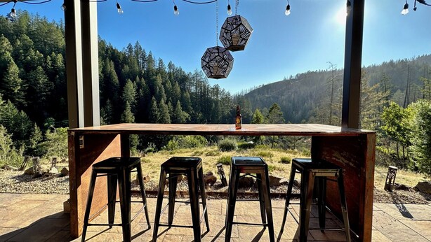 Un bar extérieur rustique, avec une table en bois et quatre tabourets, offrant une vue sur une vallée boisée.